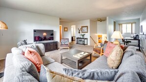 Un salon moderne avec un canapé gris, une table basse et un téléviseur. On y trouve également un coin repas avec une table et des chaises, et une cuisine à l’arrière-plan.