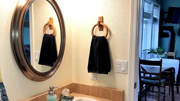 A bathroom with a round mirror, two towels hanging on wooden rings, a sink with a soap dispenser, and a window leading to a dining area. - Millers' Misfit Farm (Prineville)