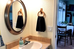 A bathroom with a round mirror, two towels hanging on wooden rings, a sink with a soap dispenser, and a window leading to a dining area. - Millers' Misfit Farm (Prineville)