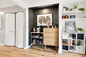 A room with a white door, a white shelving unit with a picture frame, a small wooden cabinet, and a white console table with a fan.