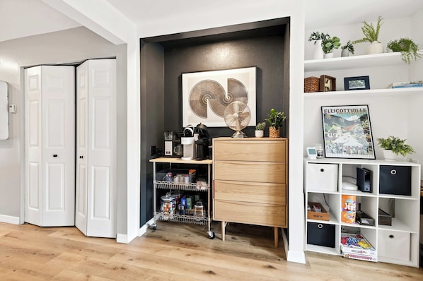 Een kamer met een witte deur, een wit wandrek met een fotolijst, een klein houten kastje en een witte console tafel met een ventilator.