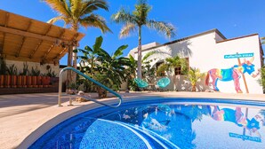 Una piscina con un diseño azul de mosaicos, rodeada de plantas tropicales y un mural de un burro colorido.
