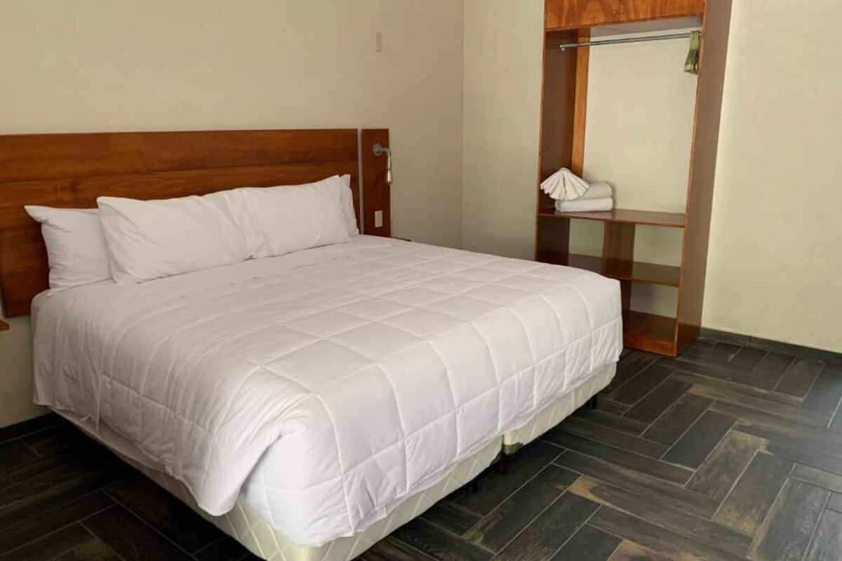 Cama bem arrumada com roupa de cama branca em quarto com piso de madeira escura e cabeceira de madeira.