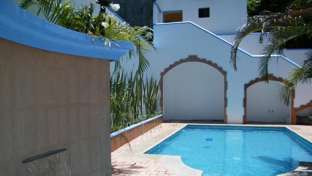 Une piscine avec un bord carrelé, un élément décoratif aquatique et une bâche bleue fixée à un mur.