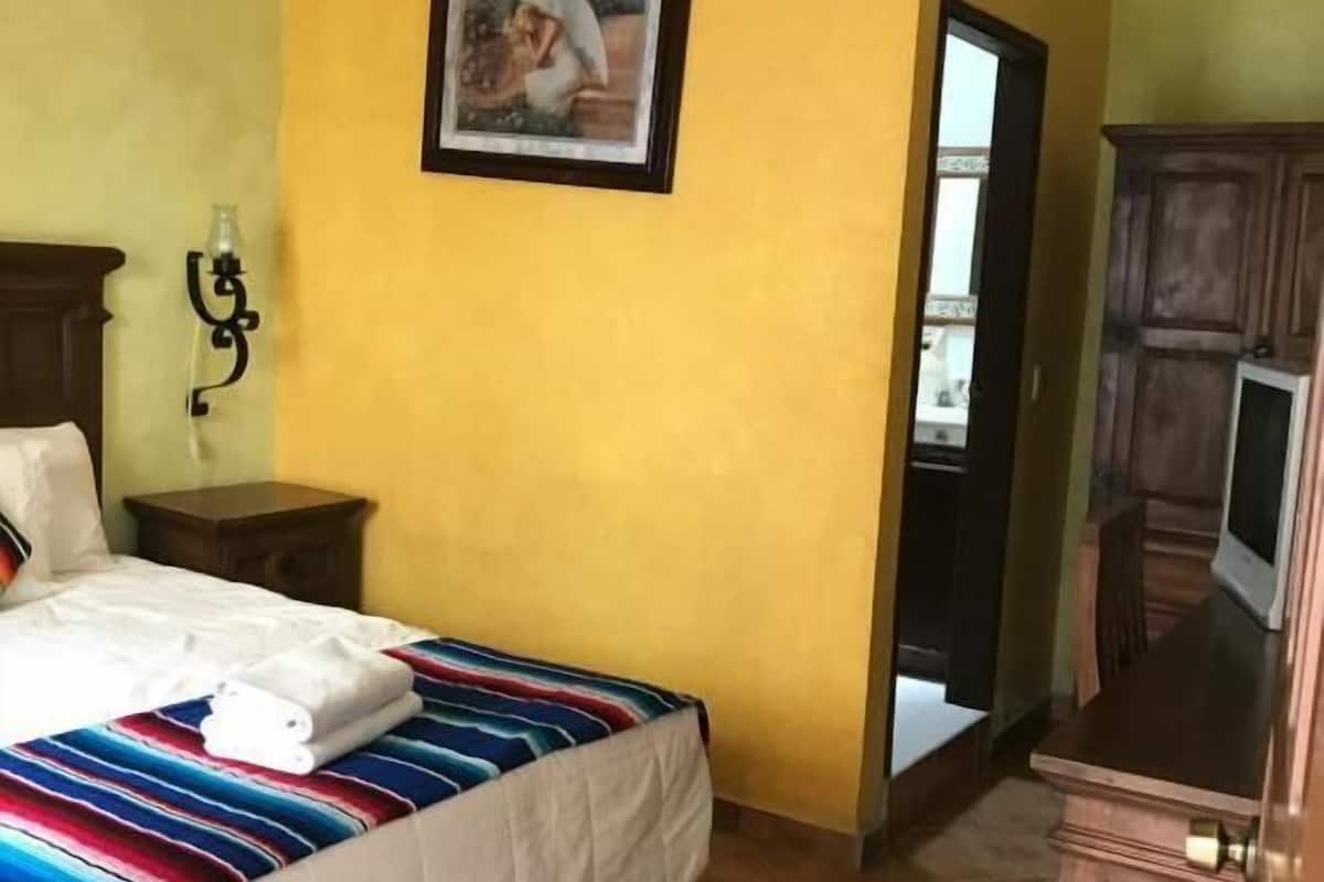 Quarto de hotel com uma cama, mesa de cabeceira, ar condicionado fixado na parede, quadro emoldurado e um armário de madeira.