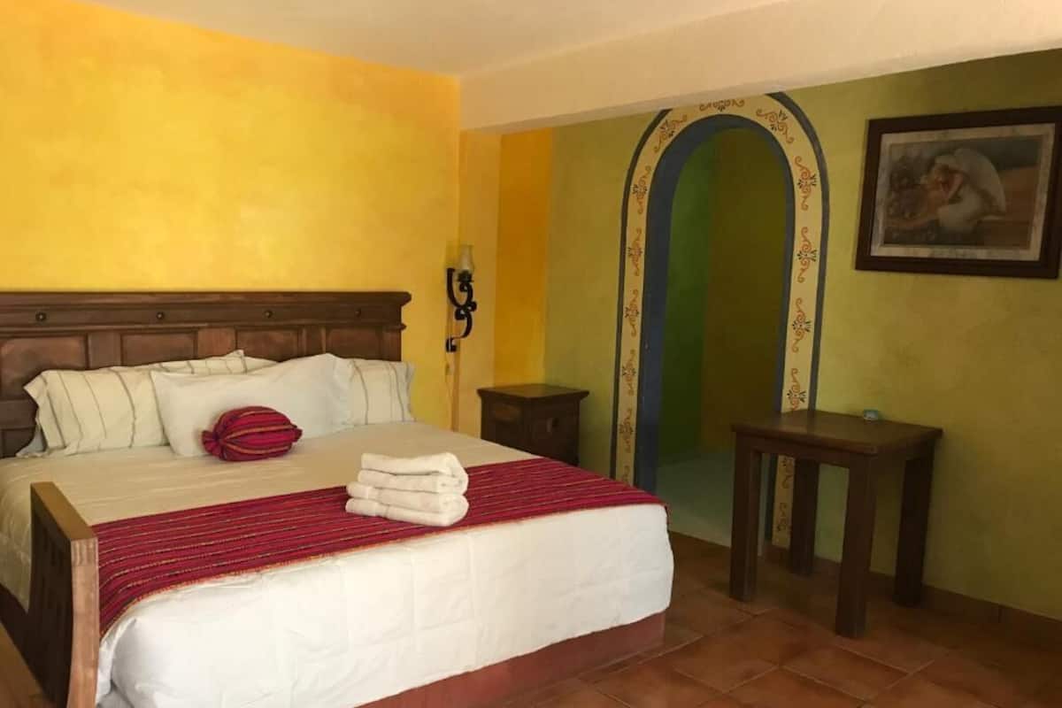 Quarto de hotel com uma cama grande, cabeceira de madeira, mesa de cabeceira pequena e um quadro na parede.
