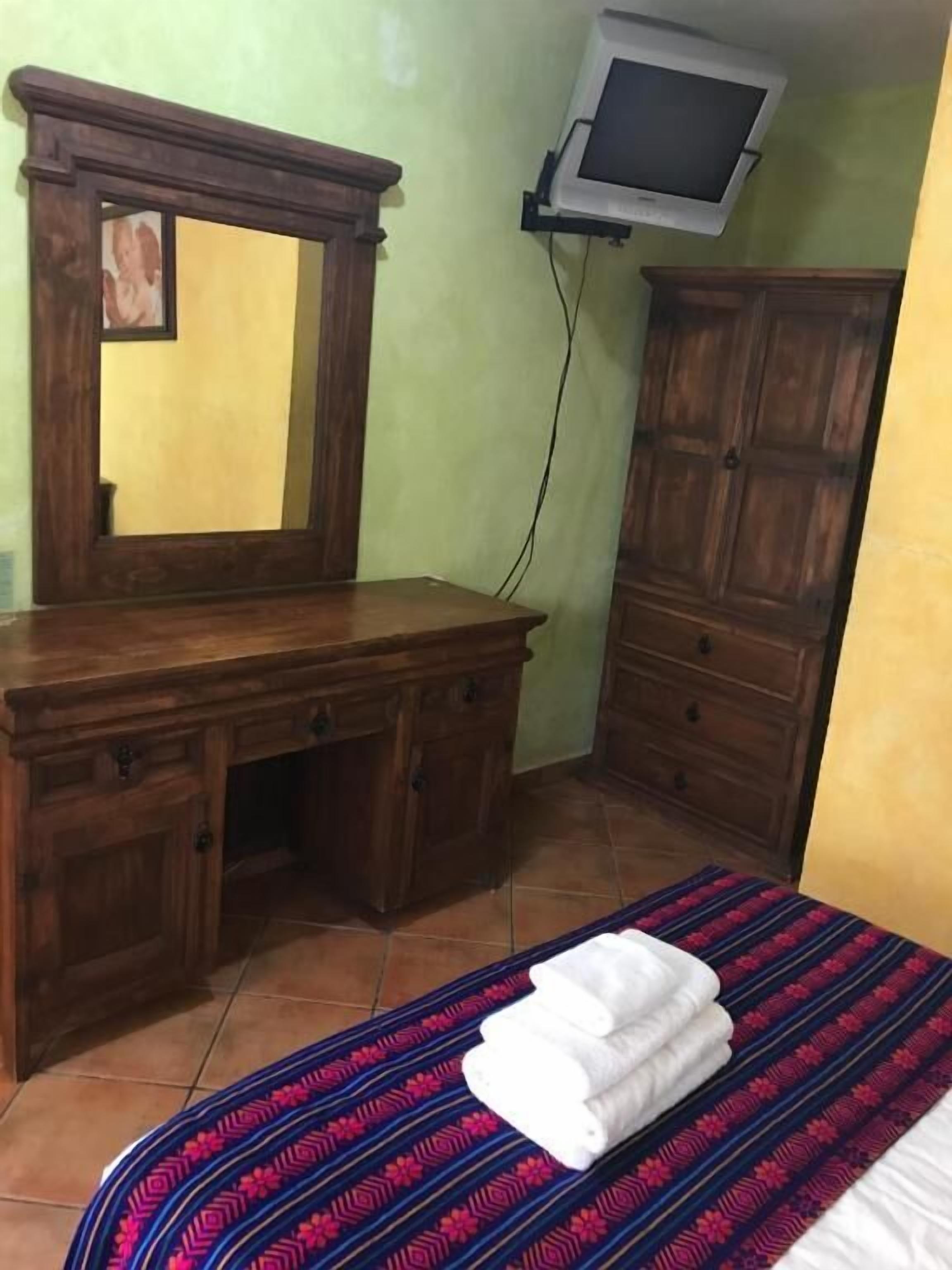 Un dormitorio con un aparador de madera, una cama con colcha estampada, un televisor montado en la pared y un espejo sobre el aparador.