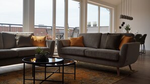 A modern living room with two sofas, a coffee table, and large windows offering a view of the cityscape. - Sleepwell Apartment - Ehrensteinsuite (Leipzig)