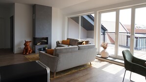 A modern living room with a grey sofa, a green armchair, and a large window offering a view of a building with a sloped roof. - Sleepwell Apartment - Ehrensteinsuite (Leipzig)