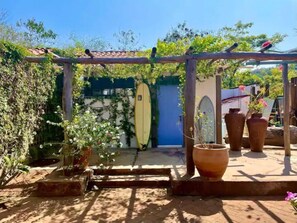 Relax in a serene outdoor space with a pergola, surrounded by lush greenery and a surfboard, perfect for those who love the ocean.