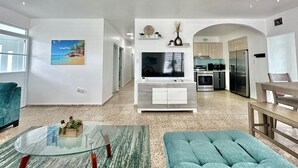 Un salón amplio con una mesa de centro de vidrio, un sofá color turquesa, un televisor de pantalla plana y una cocina visible a través de un arco.