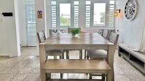 Un comedor con mesa de madera, sillas y un banco, ventanas grandes con contraventanas y un reloj en la pared.
