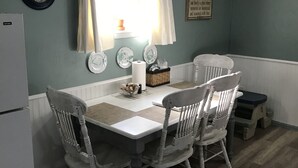 A dining area with a white table and four chairs, a clock, and a window with curtains. - Farm stay close to Colorado Springs! Pine trees, fresh air, goats and chickens! (Calhan)