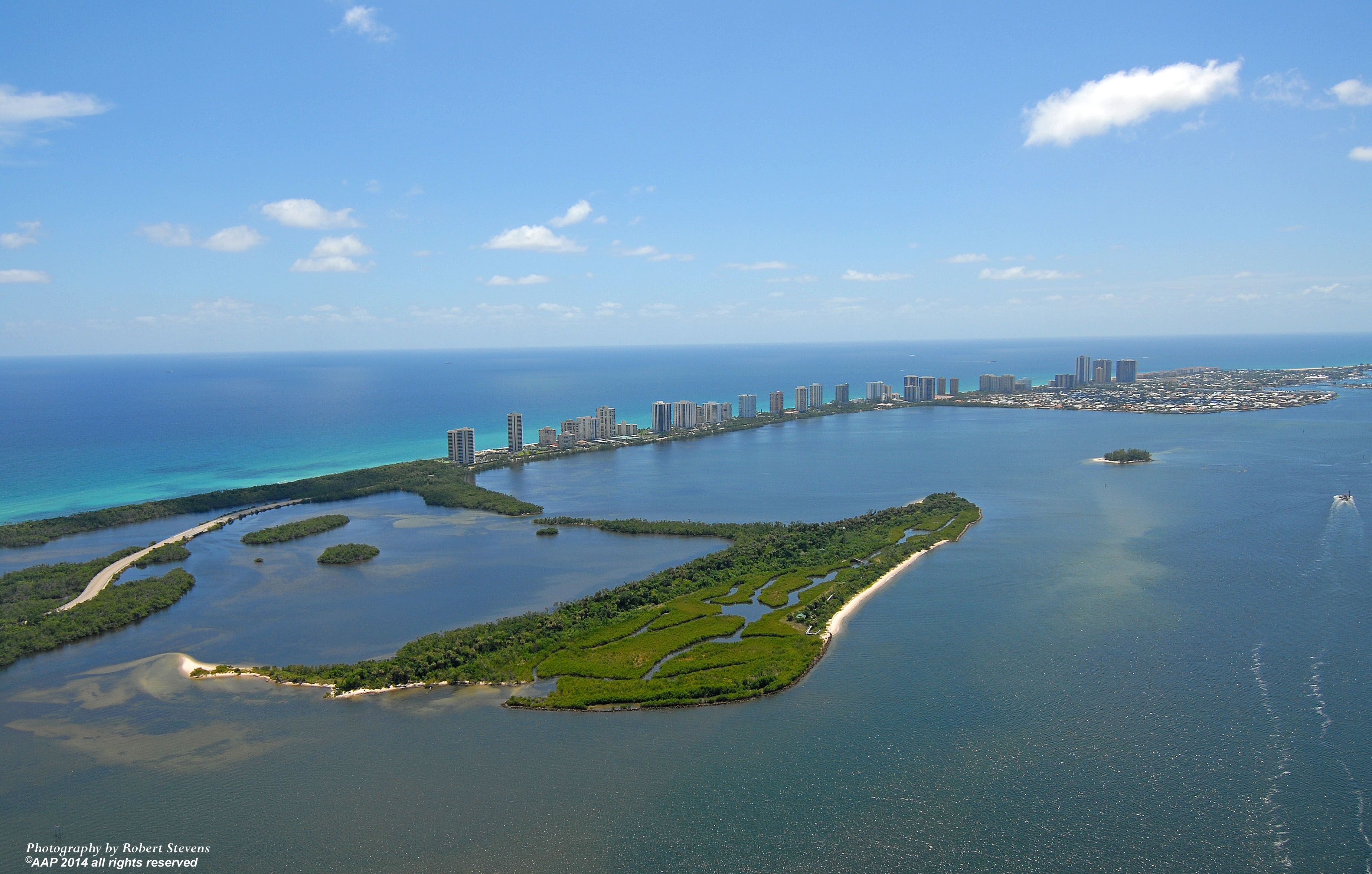 Vista aérea de una ciudad costera con un puerto deportivo, edificios altos y una extensa área verde con canales de agua.