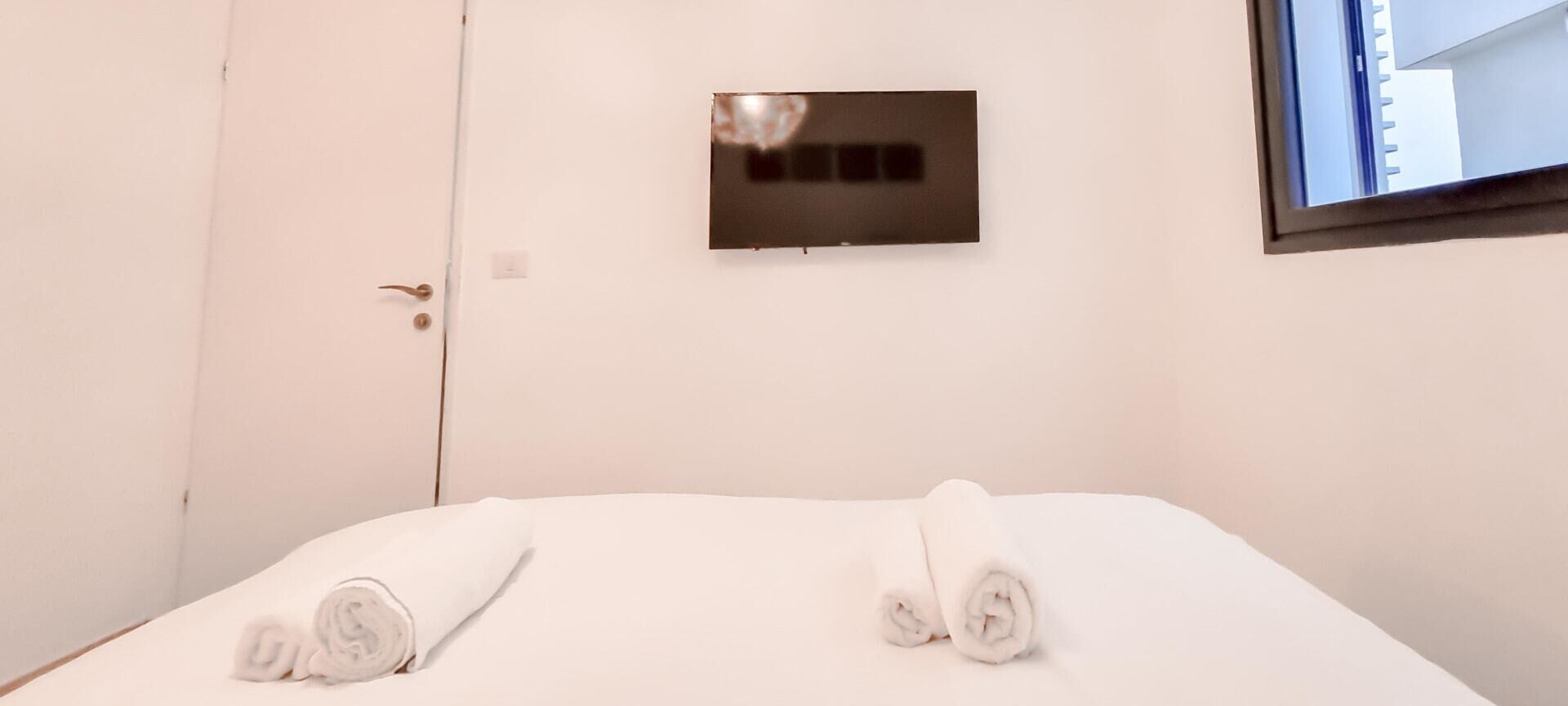 A hotel room with a bed, two rolled towels, a door, and a small wall-mounted control panel.