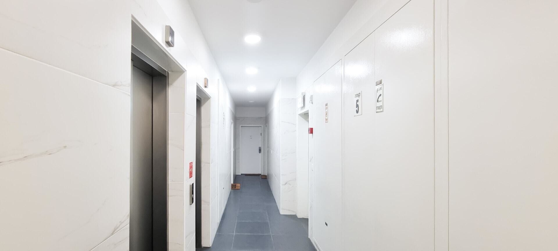 A long, narrow hallway with white walls and a grey floor, leading to a door at the end.