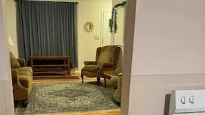 Ein schmaler Flur führt zu einem Wohnzimmer mit Sofa, Sessel und Teppich. Eine kleine Stufe führt in einen Küchenbereich mit Herd.