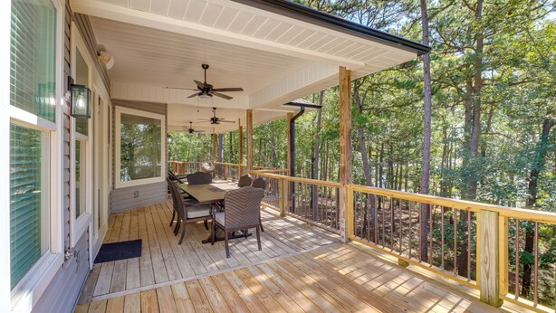 A covered deck with a dining table and chairs, surrounded by a wooden railing, overlooking a forested area. - Luxury Lakefront Retreat w/ Deck & Patio! (Edgemont)