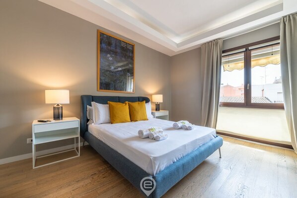 Une chambre à coucher avec un lit, deux tables de chevet, un tableau accroché au mur et une vue sur l’extérieur par une fenêtre.