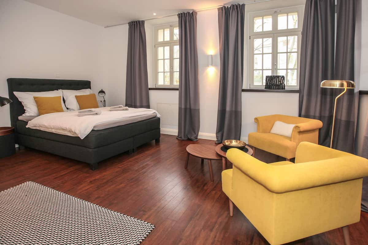 Una habitación de hotel moderna con una cama, un sillón amarillo, una mesita redonda y una lámpara de pie.