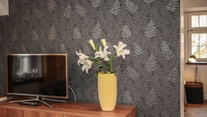 Un mueble bajo de madera para televisor con un televisor de pantalla plana, un control remoto y un jarrón amarillo con estampado que contiene flores blancas.