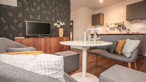Una sala de estar moderna con un sofá, un reposapiés y una mesa redonda con adornos. Un televisor de pantalla plana está montado en un mueble de madera.