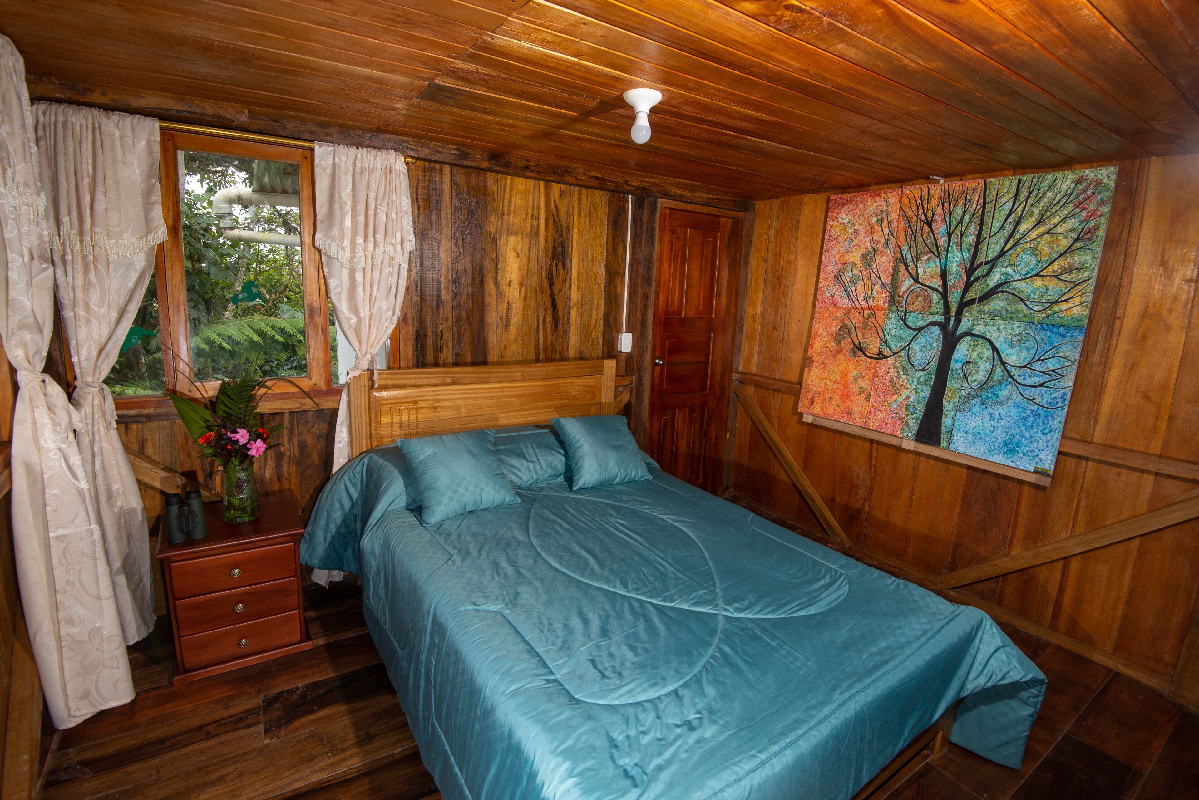 A wooden room with a bed, a nightstand, a window with curtains, and a painting of a tree.