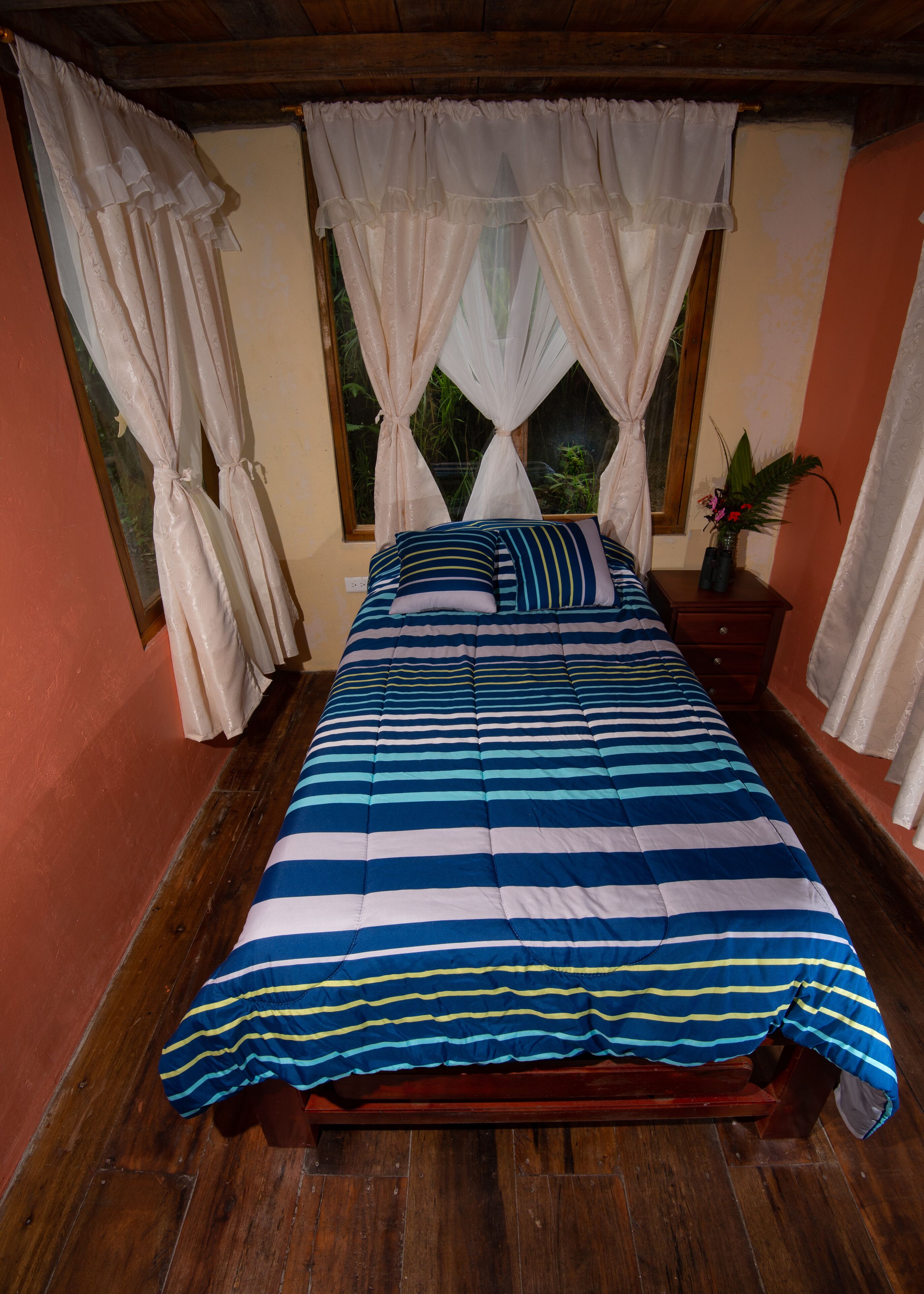 A bed with a striped blue and white bedspread, a wooden headboard, and a nightstand with a vase of flowers.