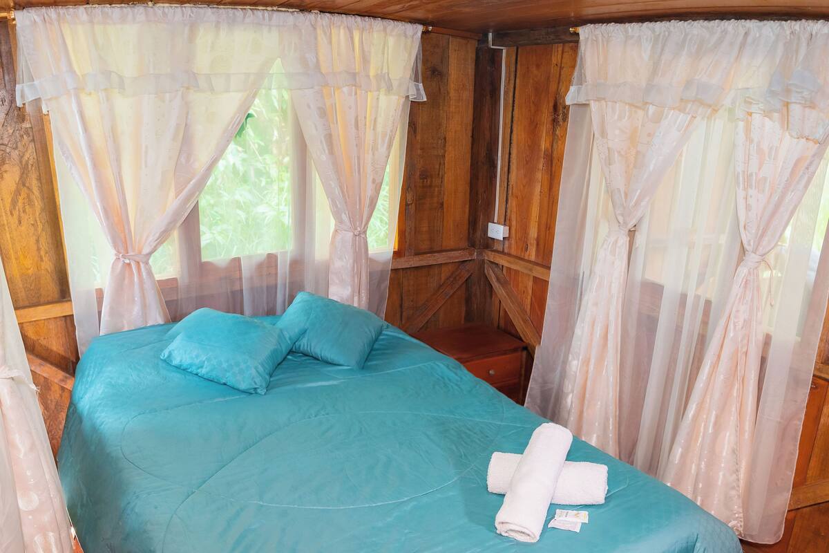 A bed with a teal bedspread and pillows, a white towel, and sheer curtains.