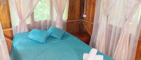 Una cama con colcha y almohadas color turquesa, una toalla blanca y cortinas de encaje.