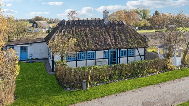 Ein traditionelles Haus mit einem Strohdach, weißer Fassade und blauen Verzierungen, umgeben von einem gepflegten Garten und einem Kiesweg.