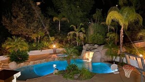 A nighttime view of a swimming pool with a waterfall feature, surrounded by tropical plants and trees. - Vacation house with spa, pool, and water slide. (El Cajon)