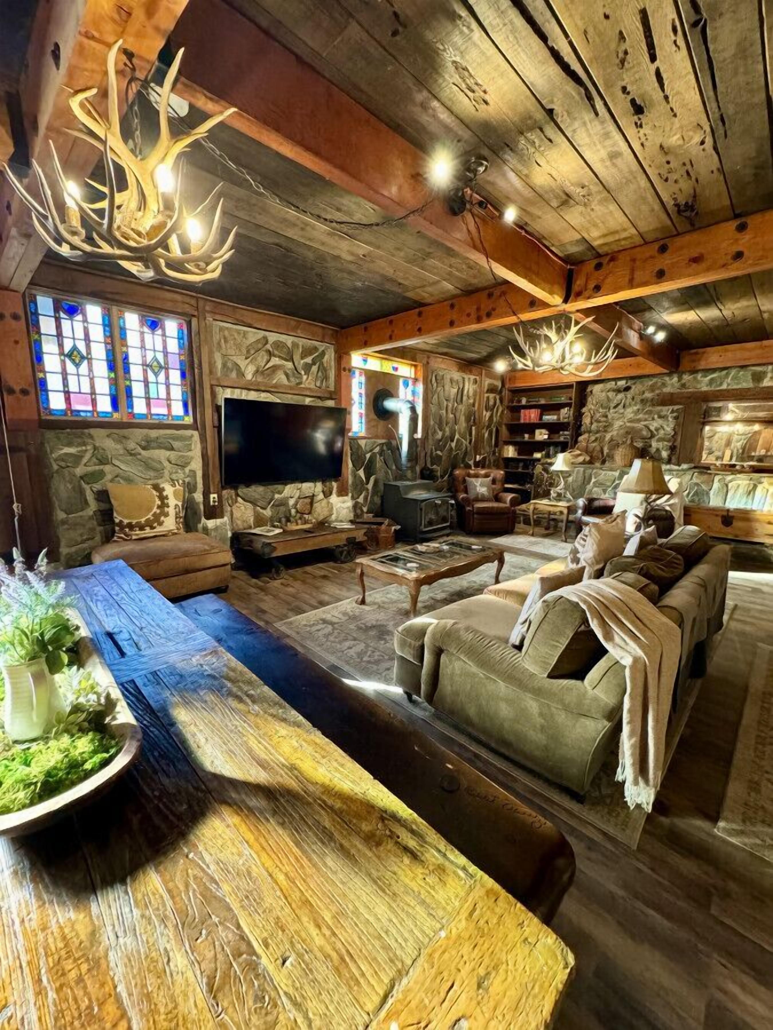 A rustic living room with a stone fireplace, wooden ceiling beams, and a large wooden dining table.