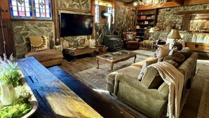 Ein rustikales Wohnzimmer mit Kamin aus Stein, Holzbalkendecke und einem großen Holztisch.
