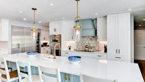 Une cuisine moderne avec des meubles blancs, un îlot central agrémenté de bols bleus et blancs, des appareils en acier inoxydable et des luminaires suspendus.