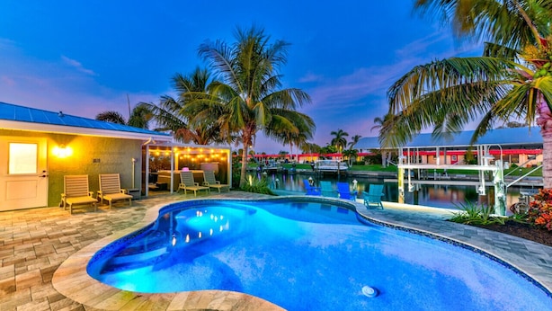 Une piscine à bordures courbes, entourée de palmiers et dotée d’une terrasse avec des chaises.