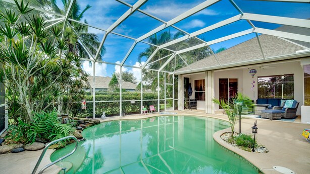 Un área de piscina con techo de vidrio, rodeada de plantas tropicales y un patio con mobiliario exterior.