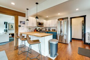 Una cocina moderna con armarios blancos, electrodomésticos de acero inoxidable, una isla de madera con taburetes altos y un cubo de basura a la vista.