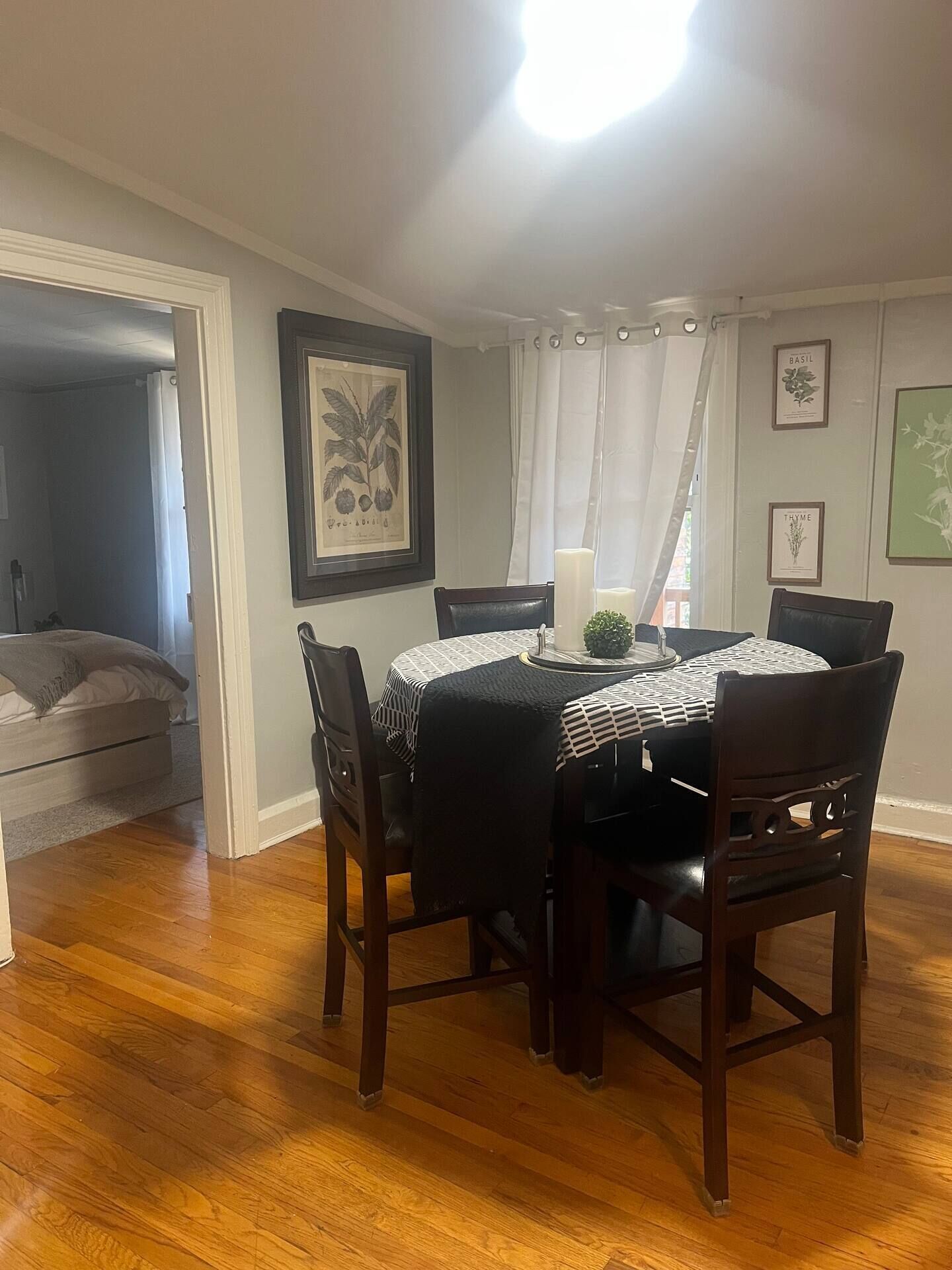 A dining area with a round table and chairs, a framed botanical print on the wall, and a large window with curtains.