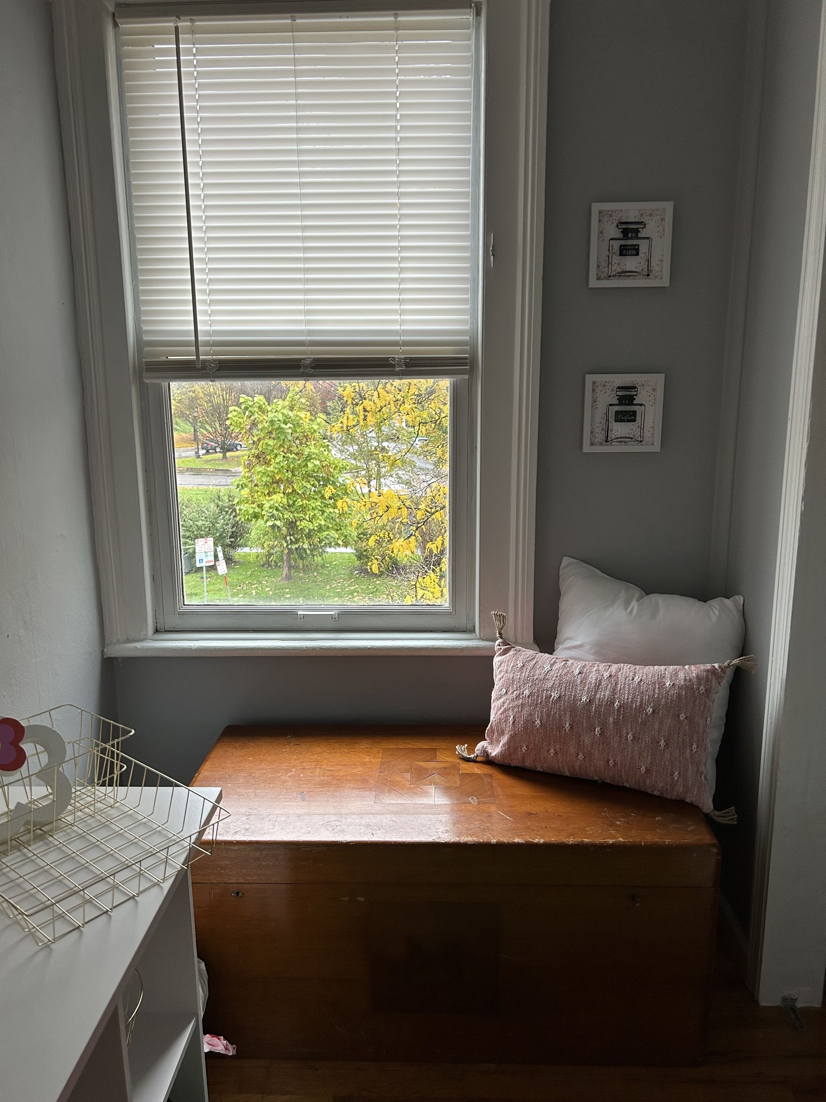A small room with a window, a wooden bench, a white pillow, and two framed pictures on the wall.