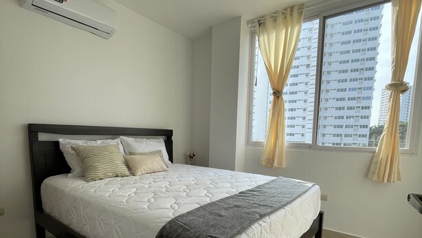 A bedroom with a bed, a window with curtains, a wall-mounted air conditioner, and a view of a building outside. - APARTAMENTOS OCEAN SUN 2 DORMITORIOS (Santa Elena)