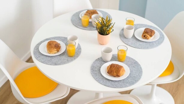 Ein Frühstückstisch mit runder Tischdecke, weiße Stühle mit gelben Kissen, Teller mit Croissants, Gläser Orangensaft und ein kleiner Blumentopf.