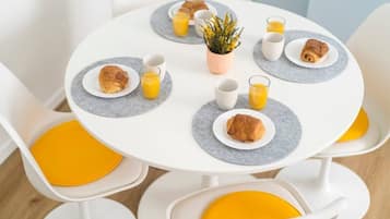 A breakfast setting with a round table, white chairs with yellow cushions, plates with croissants, glasses of orange juice, and a small potted plant.