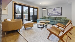 Un salon moderne comprenant un canapé, des fauteuils, une table basse et une grande fenêtre donnant sur des arbres et un immeuble.