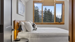 Une chambre à coucher avec un lit, une table de chevet avec une lampe, une fenêtre donnant sur des arbres et des montagnes, et un tableau accroché au mur.
