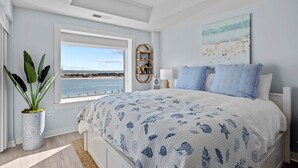 Un dormitorio con una cama grande, vista al mar, una planta en maceta y una estantería.