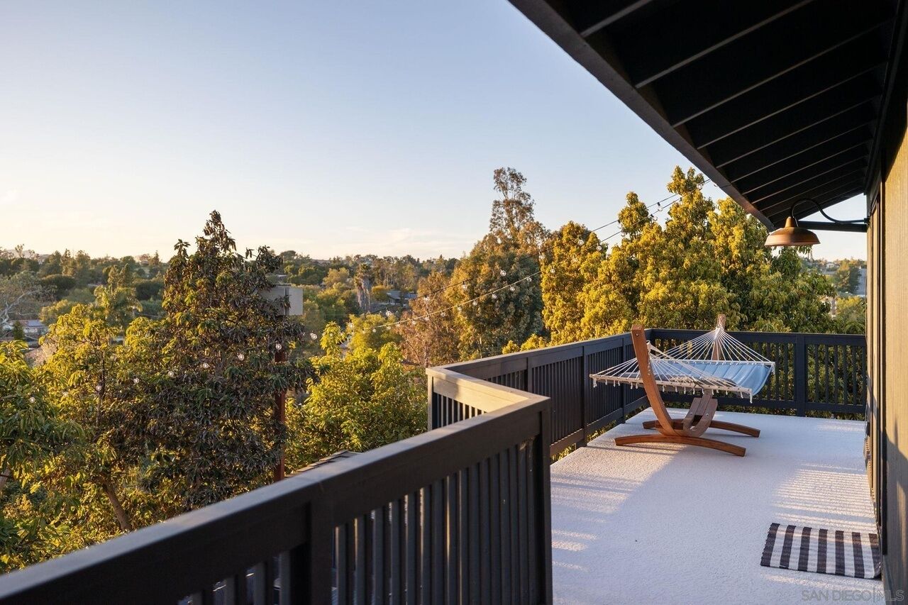 A balcony with a hammock chair, a view of trees and buildings, and a clear sky.
