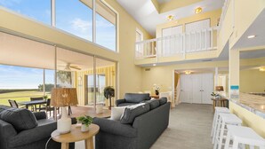 A spacious living area with a grey sofa, a round wooden table, and a small potted plant. There is a balcony with a railing and a view of the outdoors. - One-of-a-Kind Villa in Beach Castle! Direct Views of the Bay, Walk to Beach (Longboat Key)