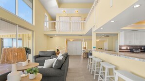 A spacious living area with a grey sofa, a wooden table with a lamp, and a kitchen area with white bar stools. - One-of-a-Kind Villa in Beach Castle! Direct Views of the Bay, Walk to Beach (Longboat Key)