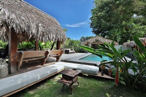 A thatched-roof hut with a wooden bench and a small table, a pool, and lush greenery.